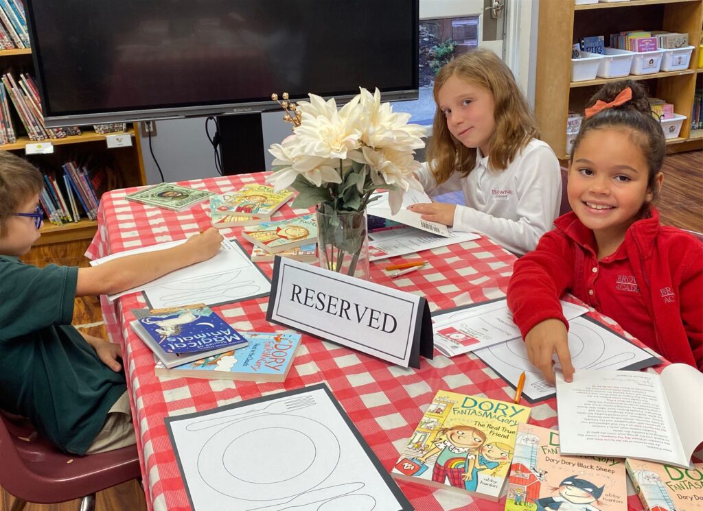 3 students sit at a table for reading exercises, two are smiling at the camera for a photo