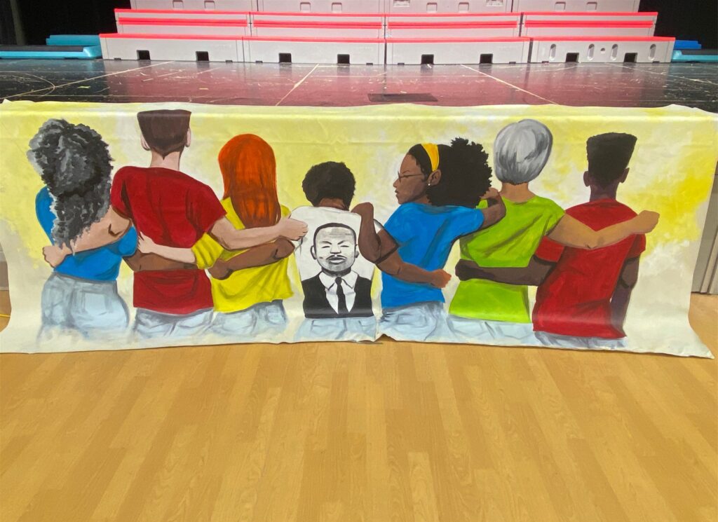 A poster illustrating a diverse group of people ,arm-in-arm, with their backs to the viewer and the middle person's shirt has an illustration of MLK Jr.
