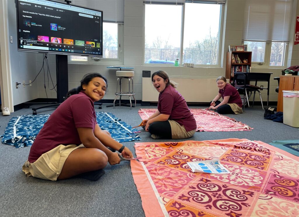 Three students sit cutting fabric for blankets, smiling at the camera