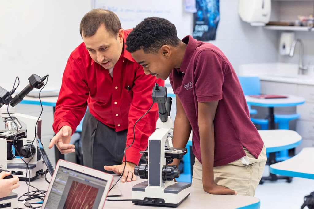 a science teacher and student confer over a microscope