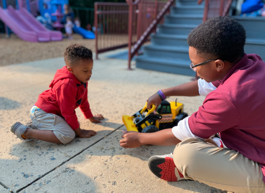 An older students and younger student sit outside playing with a toy truck