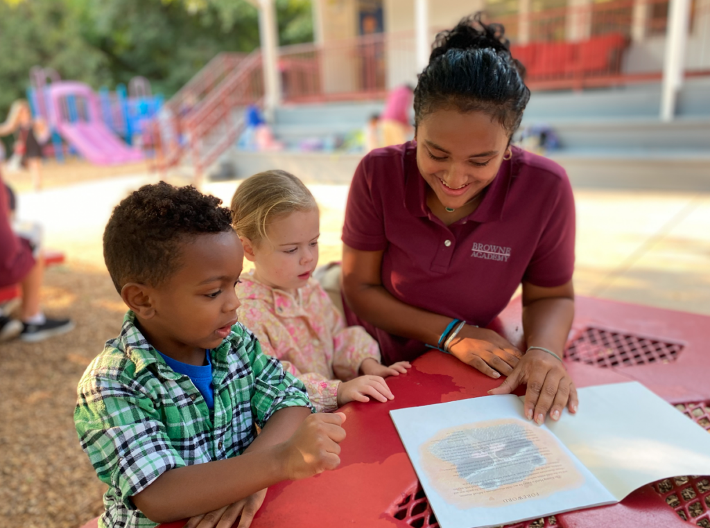 An older student sits outside with two younger students reading a book to them