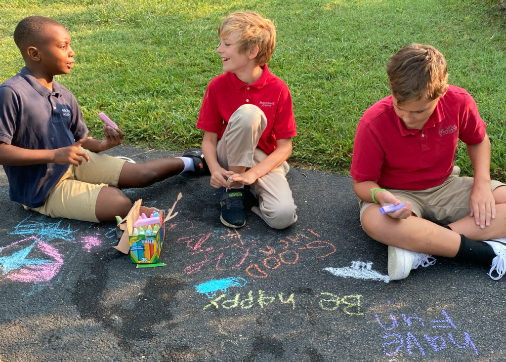 Three students sit outside drawing chalk, two of them are laughing together