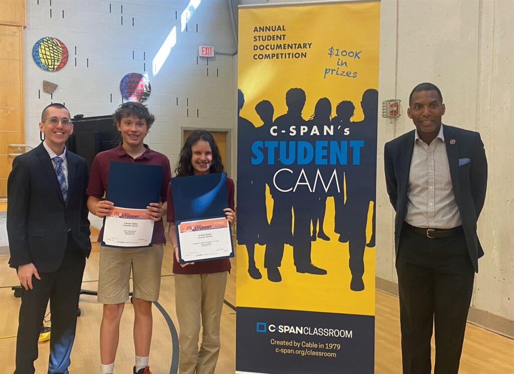 Two students and two event organizers stand for a photo next to a 'C-SPAN Student Cam' event sign