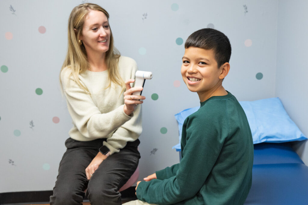 A School nurse holds a thermometer out to a student who is sitting on the examination table and smiling at the camera